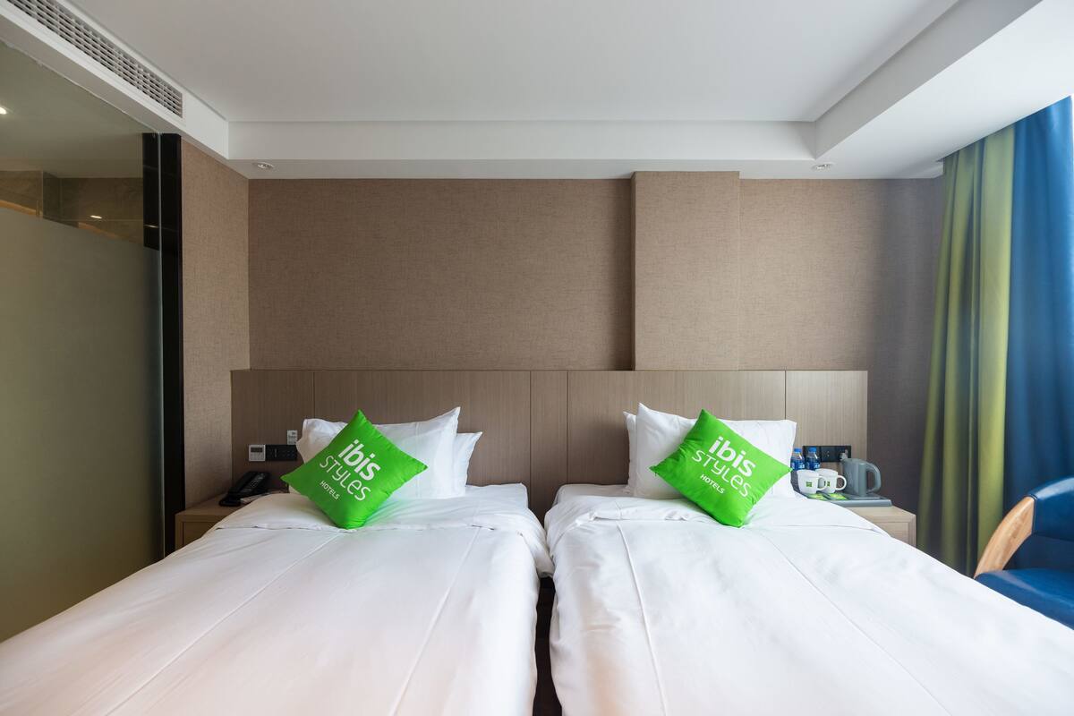 Habitación de hotel con dos camas, cada una con ropa blanca y almohadas verdes. Hay una mesita de noche con un teléfono y una cafetera.