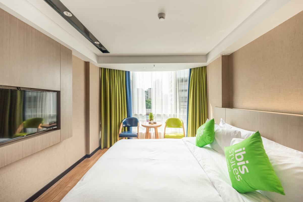 Habitación de hotel con cama, televisor, una mesita con sillas y balcón con cortinas.
