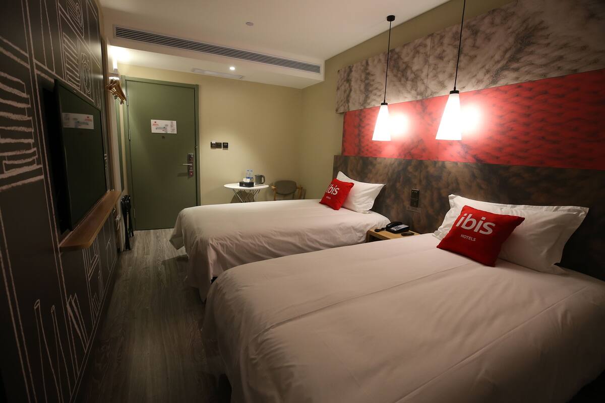 A hotel room with two beds, each with a red pillow labeled "ibis Hotels," a small table with a coffee maker, and a chair.