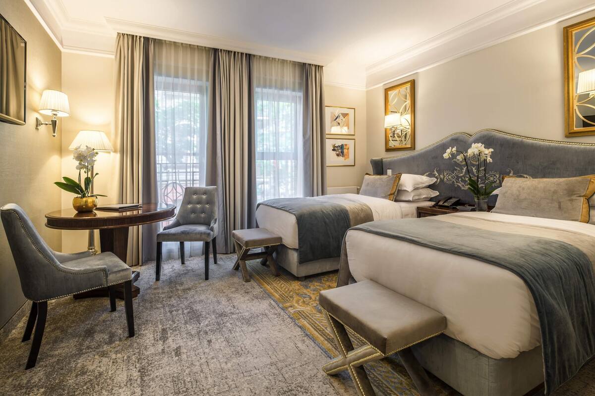 A hotel room with a bed, a chair, a small table with a lamp, and a window with curtains.