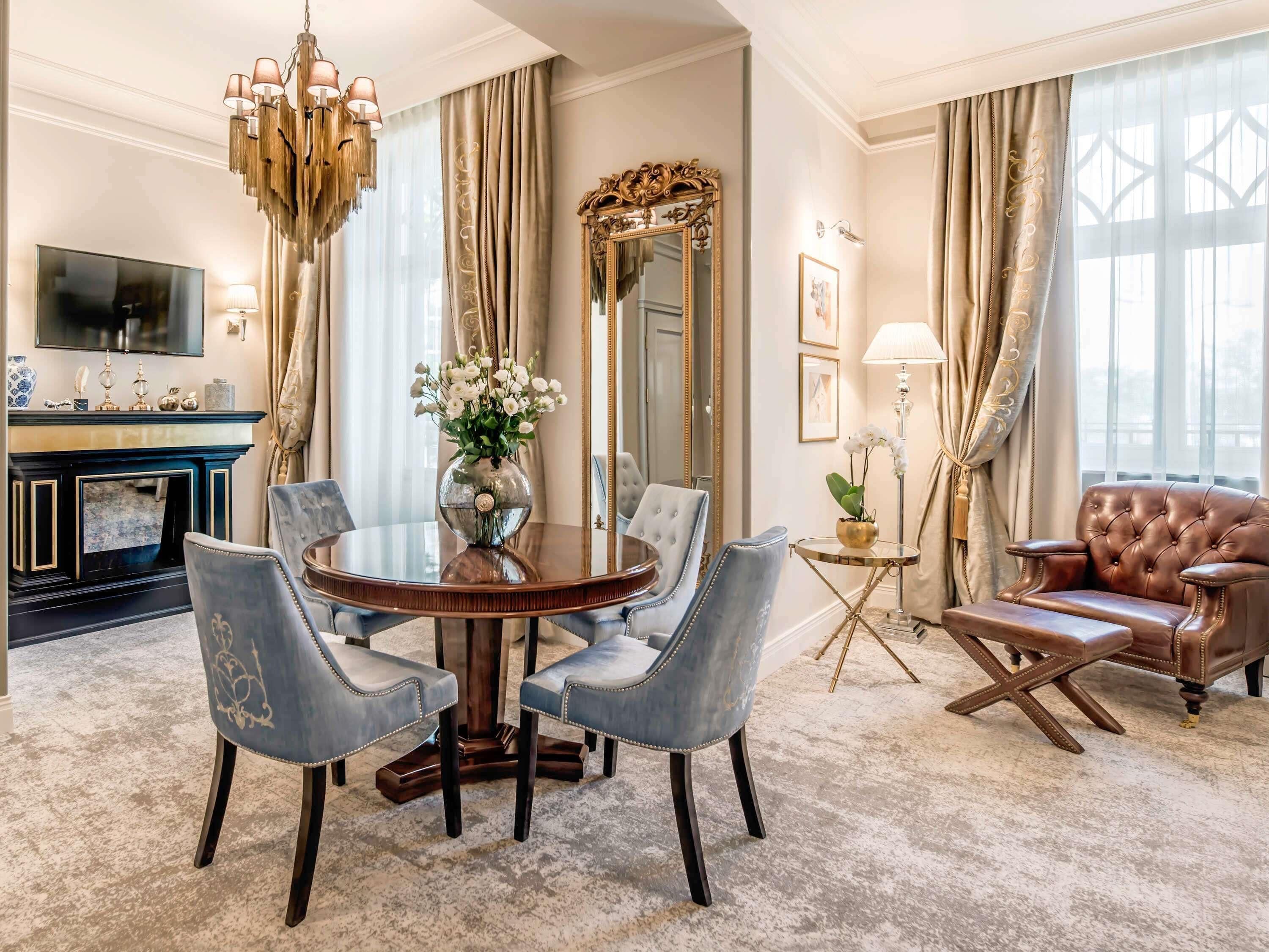 A formal dining room with a round table, chairs, a chandelier, a fireplace, a large mirror, and a leather armchair.