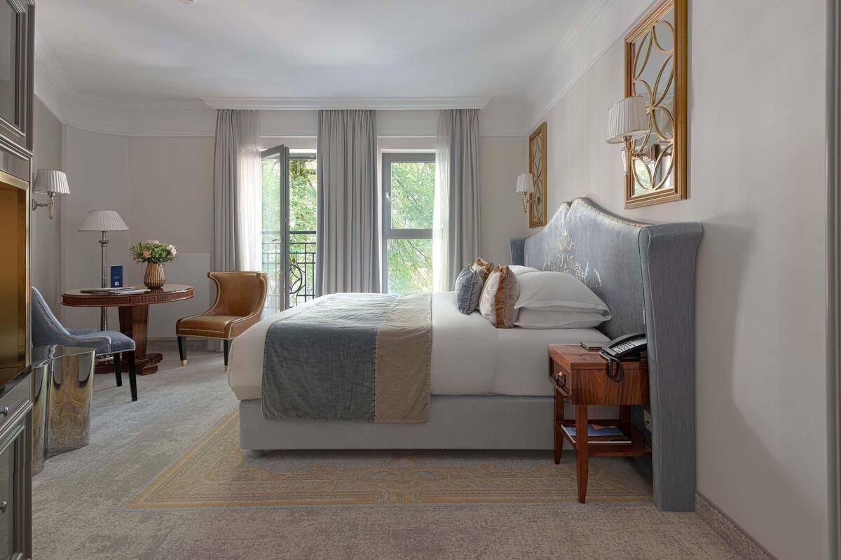 A hotel room with a large bed, a desk with a lamp, a chair, and a view of greenery through the window.