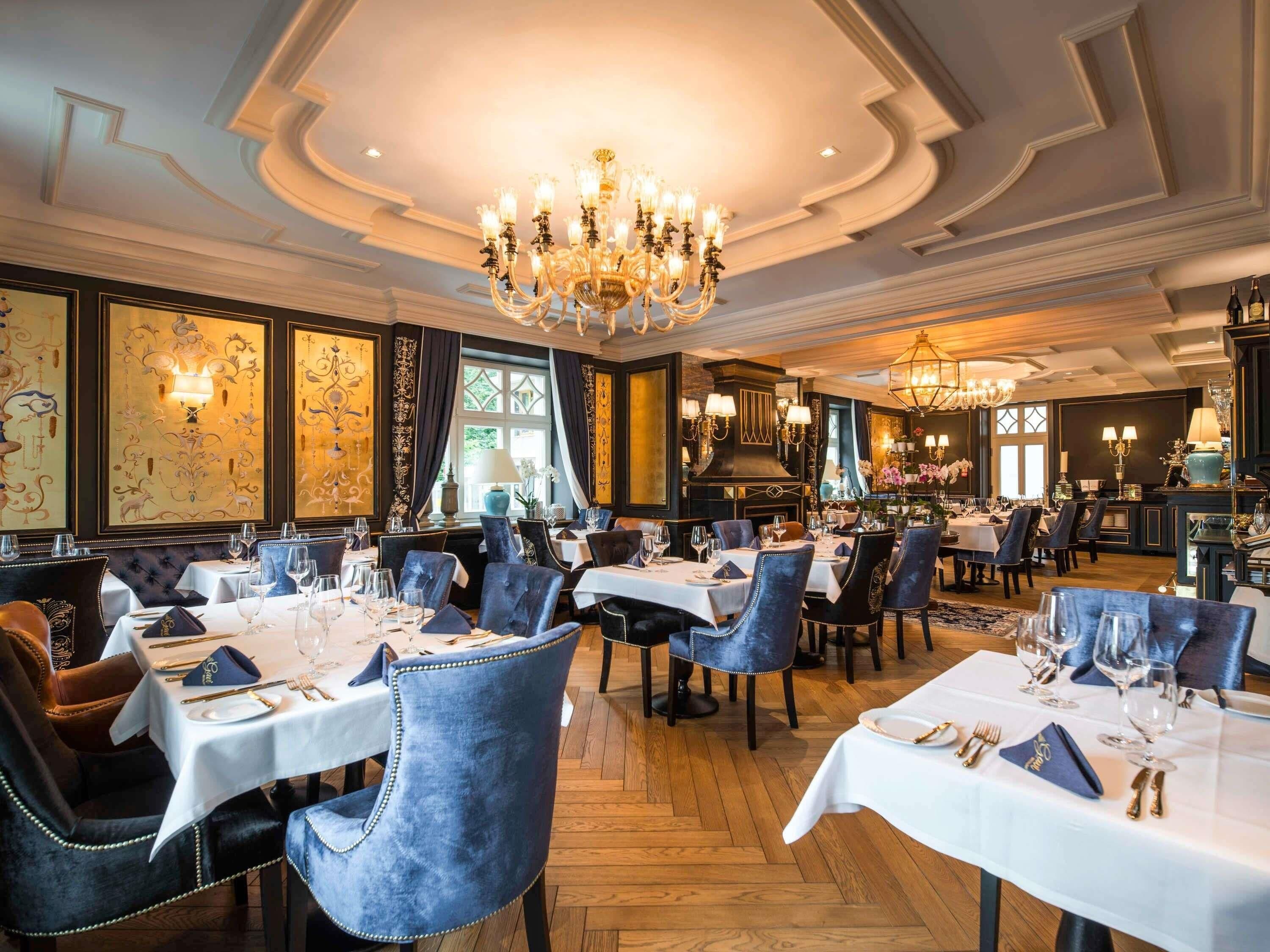 A formal dining room with multiple tables, elegant chairs, chandeliers, and decorative wall panels.