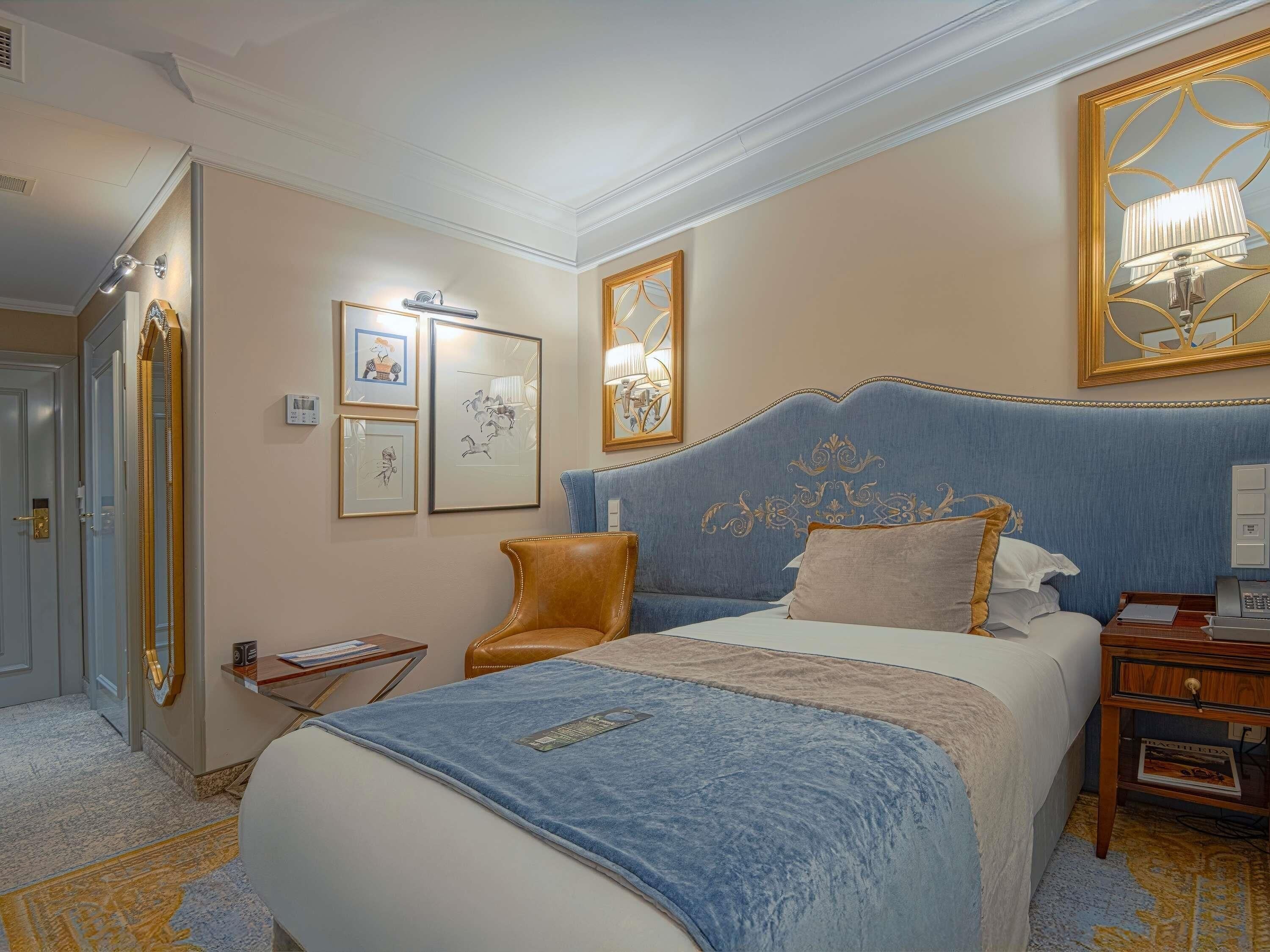 A hotel room with a large bed, a blue headboard, a brown leather chair, and a nightstand with a lamp.