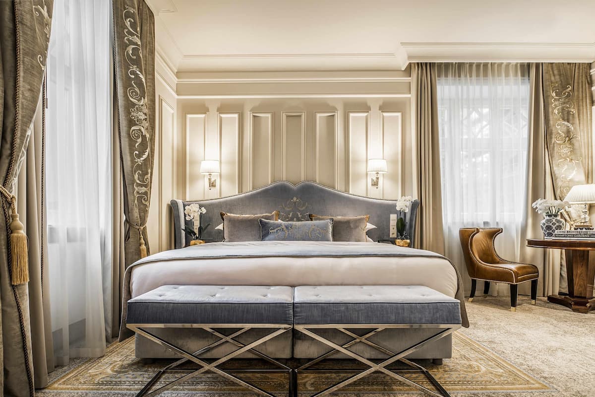 A large bed with a tufted headboard, a bedside table with a lamp, and a chair in a hotel room with a patterned carpet.