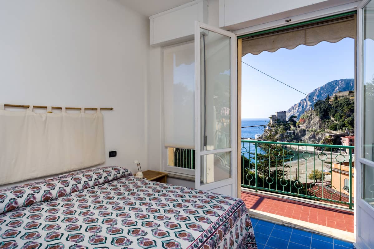 A bedroom with a bed, a window, and a balcony with a view of the sea and buildings.