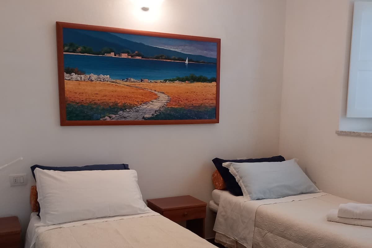 A hotel room with two beds, each with white linens and pillows, a painting of a coastal landscape above the beds, and a window on the right side.