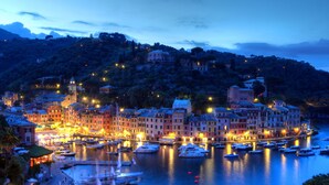 Une ville côtière au crépuscule, avec des bâtiments illuminés, un port de plaisance rempli de bateaux et une colline en arrière-plan.