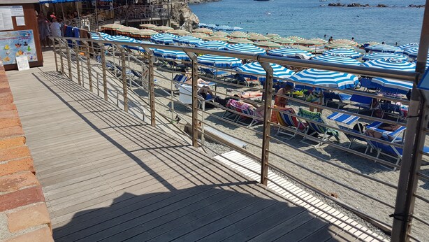 Une terrasse au bord de la plage, bordée de rangées de parasols à rayures bleues et blanches, une promenade en bois et, au loin, une côte rocheuse.