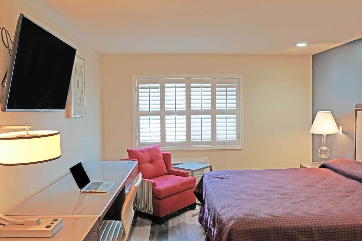 A hotel room with a bed, a desk with a laptop, a chair, a TV mounted on the wall, and a window with blinds.