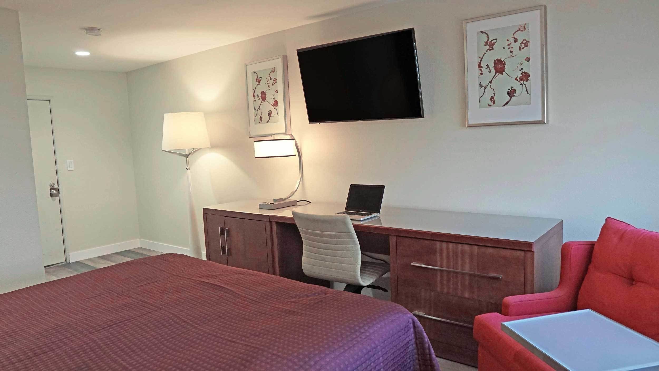 A hotel room with a bed, a desk with a computer, a chair, a TV, and artwork on the walls.