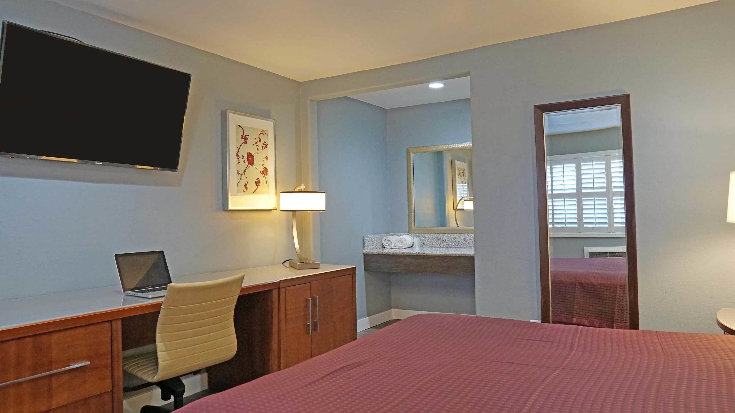 A hotel room with a bed, a desk with a laptop, a chair, a TV mounted on the wall, a framed picture, and a mirror.