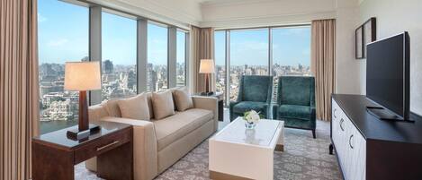 Habitación de hotel moderna con un amplio ventanal que ofrece vistas a la ciudad, un sofá, una mesa de centro, un televisor y un armario.
