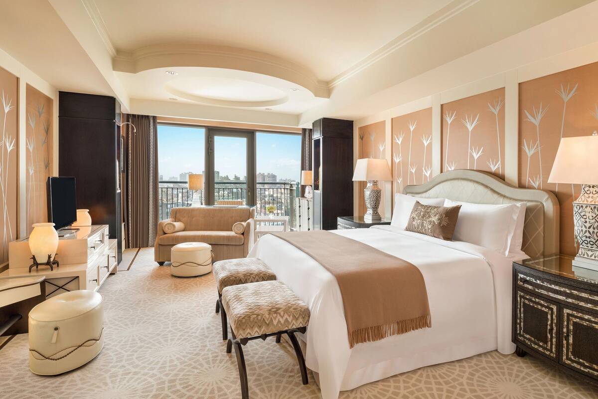 Amplia habitación de hotel con una cama grande, una zona de estar con un sillón y reposapiés, un escritorio con lámpara y vistas a la ciudad a través de amplios ventanales.