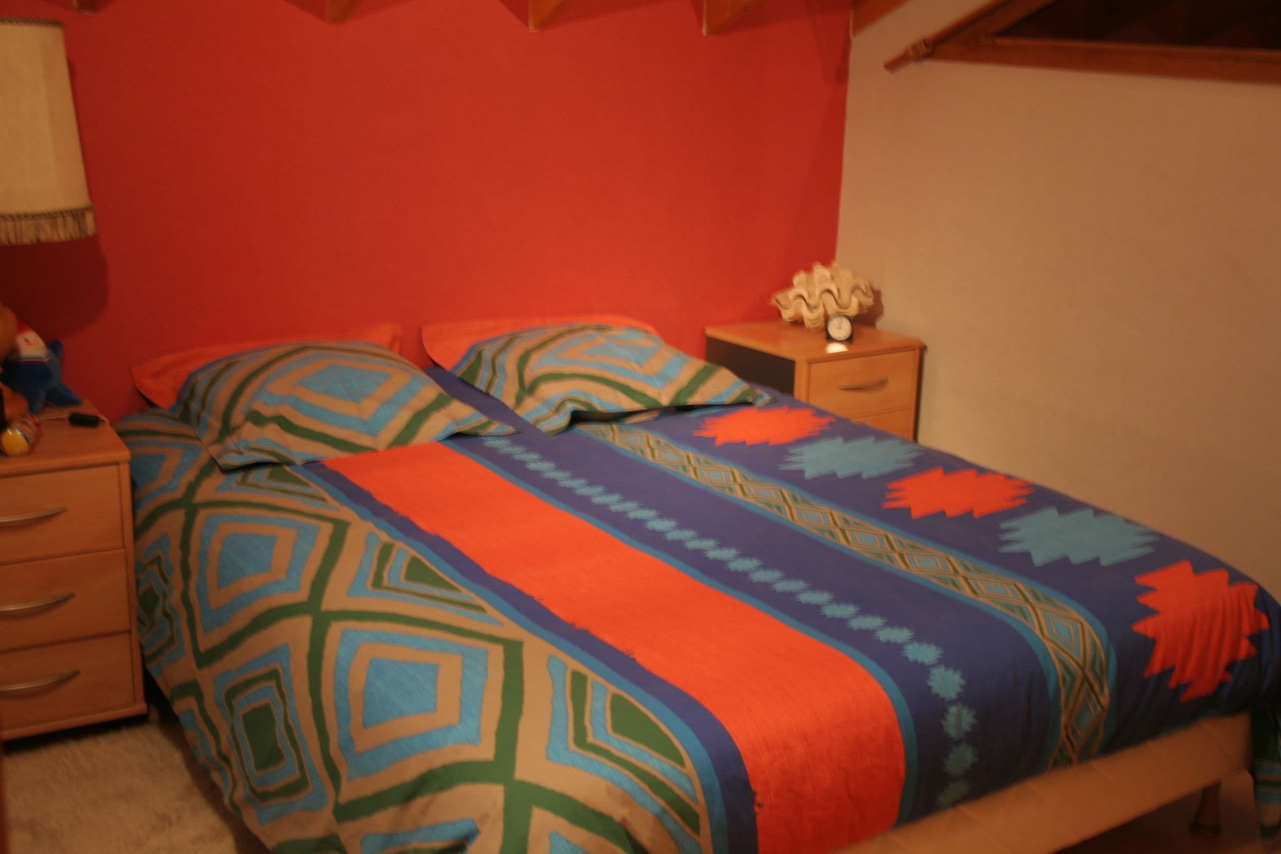 A bedroom with a bed covered in colorful bedding, a nightstand with a lamp, and a dresser.