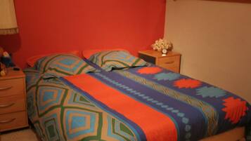 A bedroom with a bed covered in colorful bedding, a nightstand with a lamp, and a dresser.