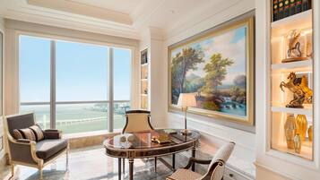 Um quarto luxuoso com uma grande janela que oferece vista para a cidade, piso de mármore, uma mesa redonda com luminária e uma estante com objetos decorativos.