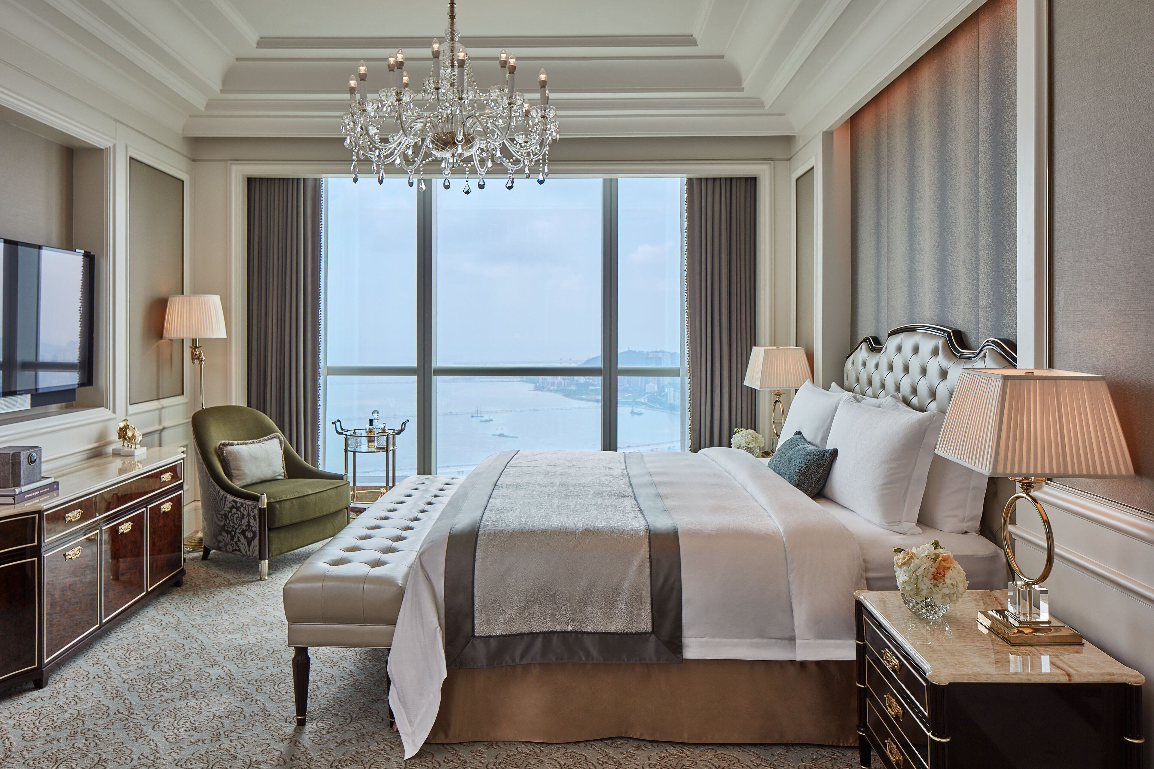 Amplia habitación de hotel con una cama grande, televisión, candelabro y vistas de la ciudad a través de amplias ventanas.