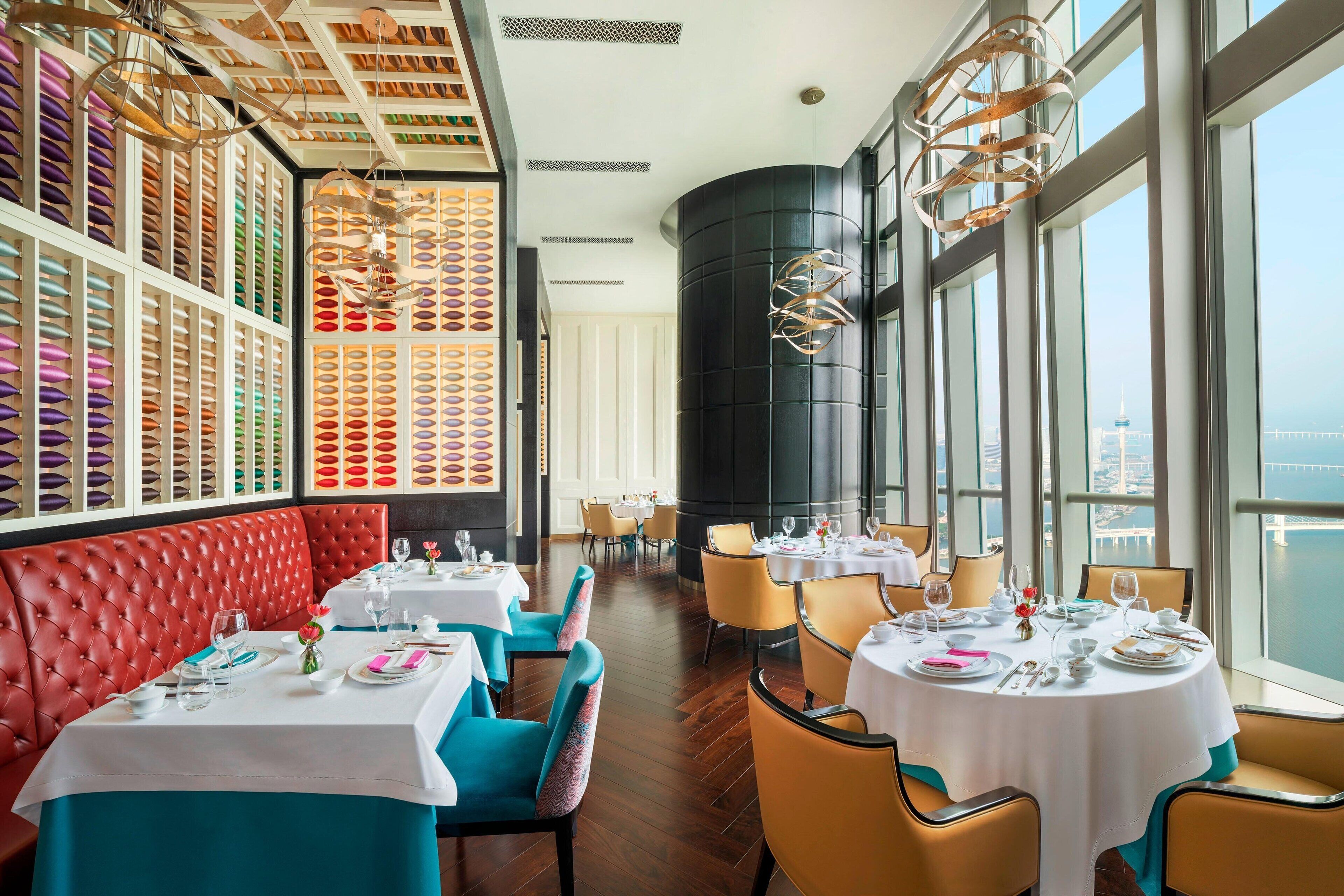 Ein Restaurant mit roten Ledersesseln, türkisfarbenen Stühlen und weißen Tischen. Große Fenster bieten einen Blick auf die Stadt.