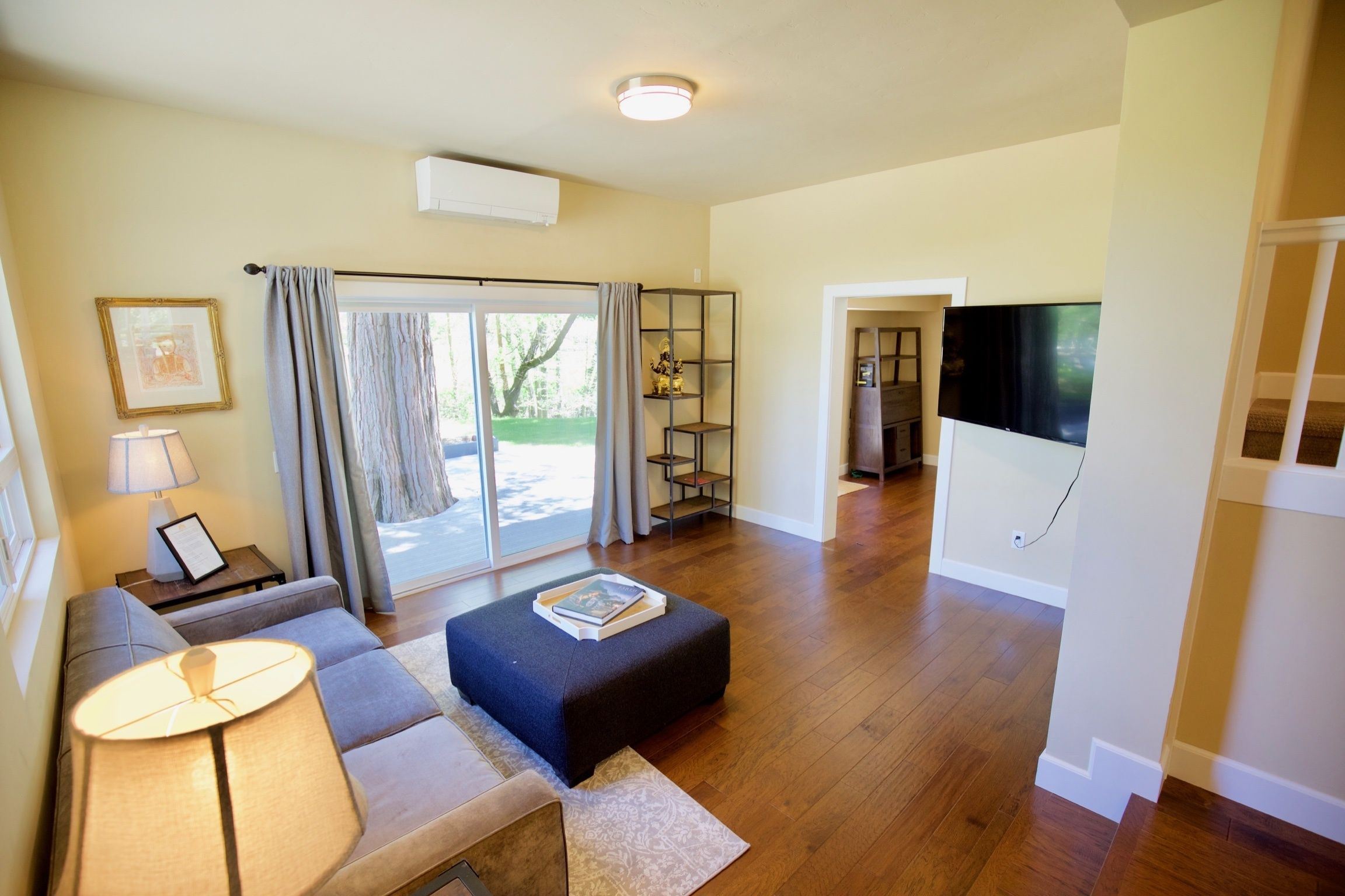 Sala de estar espaçosa com sofá, banquinho e luminária. Possui uma porta de vidro deslizante que dá para uma área externa.