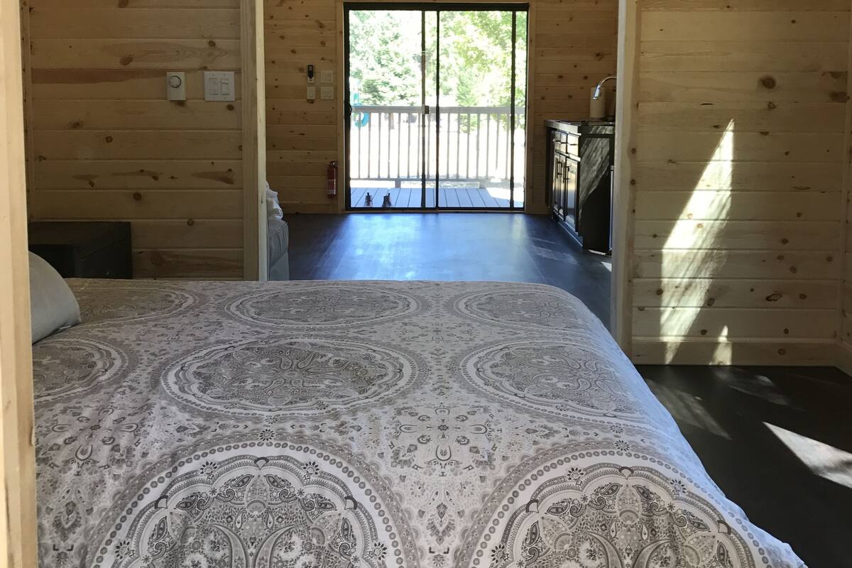 A wooden cabin room with a bed, a small table, and a view of a deck through a large window.