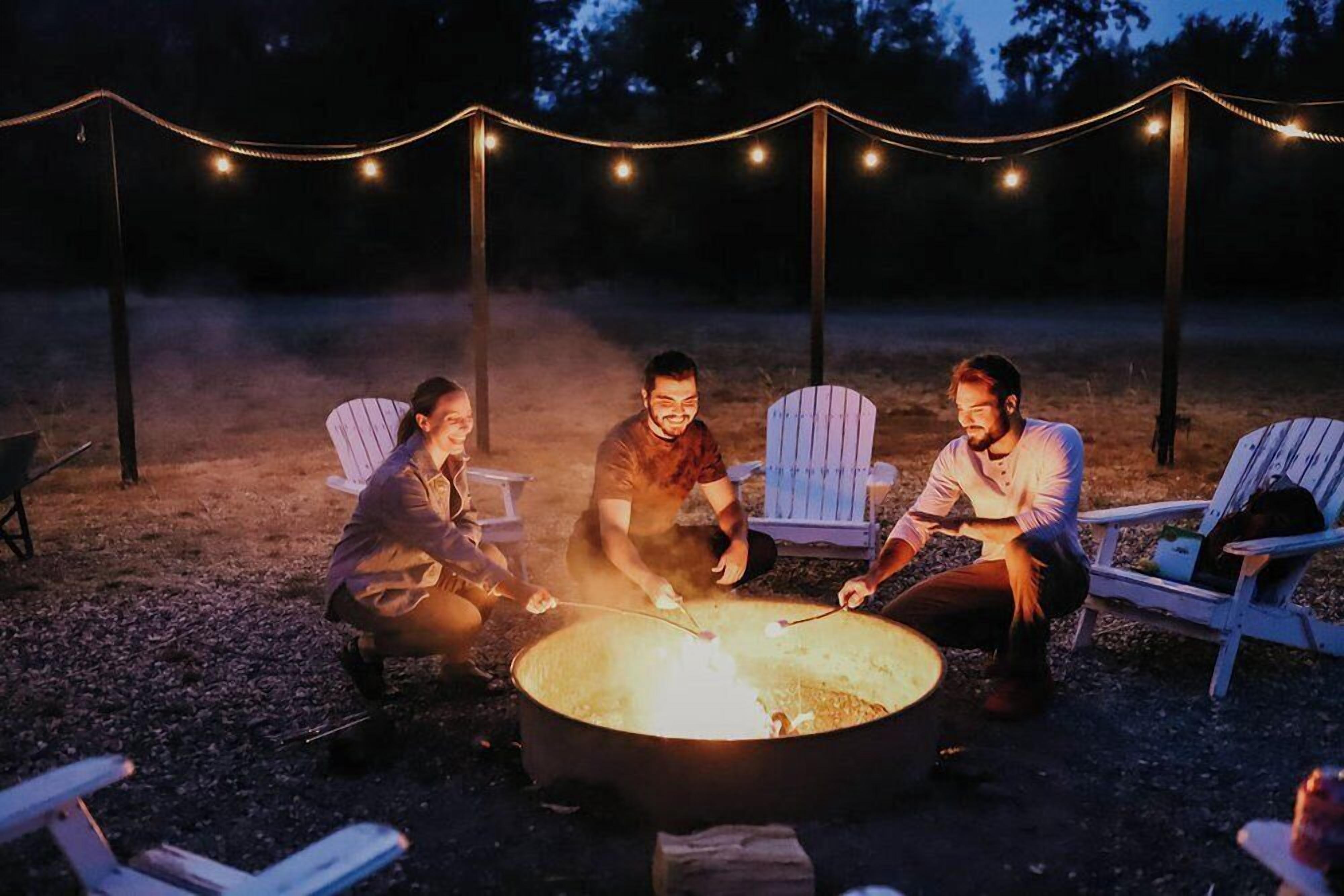 Three people sitting around a campfire with chairs and string lights in the background.
