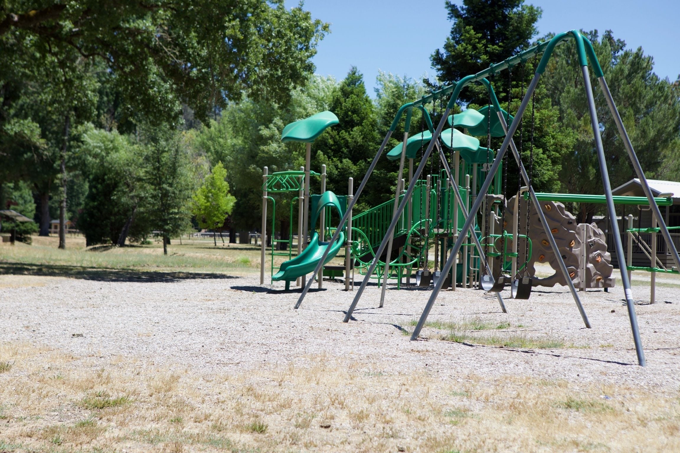 Un parque infantil con un tobogán y estructuras para escalar, rodeado de árboles y suelo de grava.