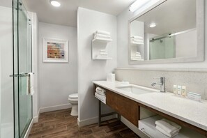 A modern bathroom with a glass shower enclosure, a large mirror, and a wooden vanity with a sink and towel storage. - Hampton Inn & Suites Johns Creek (Johns Creek)