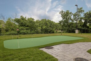 Um campo de mini golfe com uma área de putting verde, uma cerca de metal preto e um caminho pavimentado.