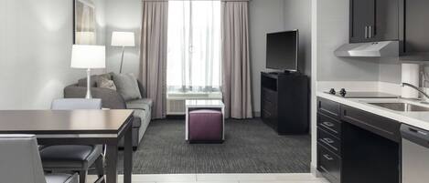 A modern hotel room with a dining area, a sofa, a TV, and a kitchen.