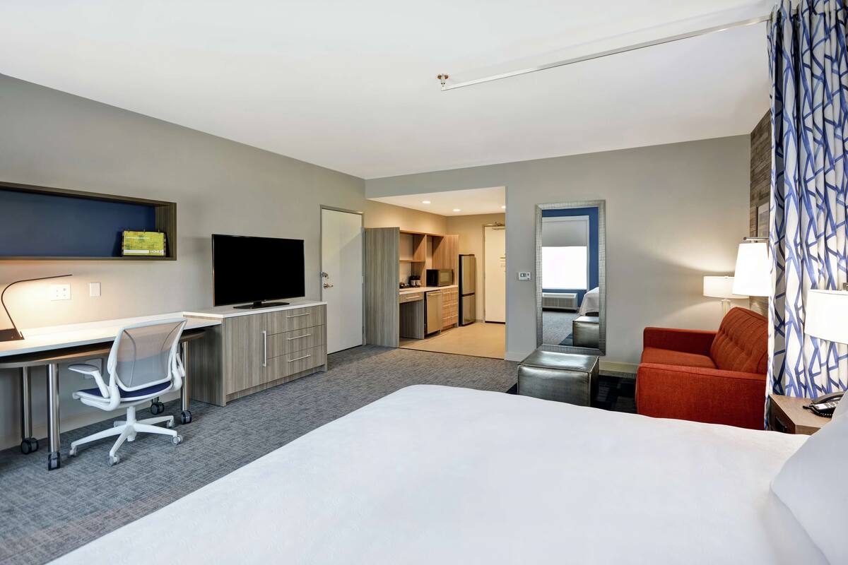A hotel room with a bed, desk, chair, TV, kitchenette, and a bathroom with a mirror and towel rack.