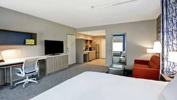 A hotel room with a bed, desk, chair, TV, kitchenette, and a bathroom with a mirror and towel rack.