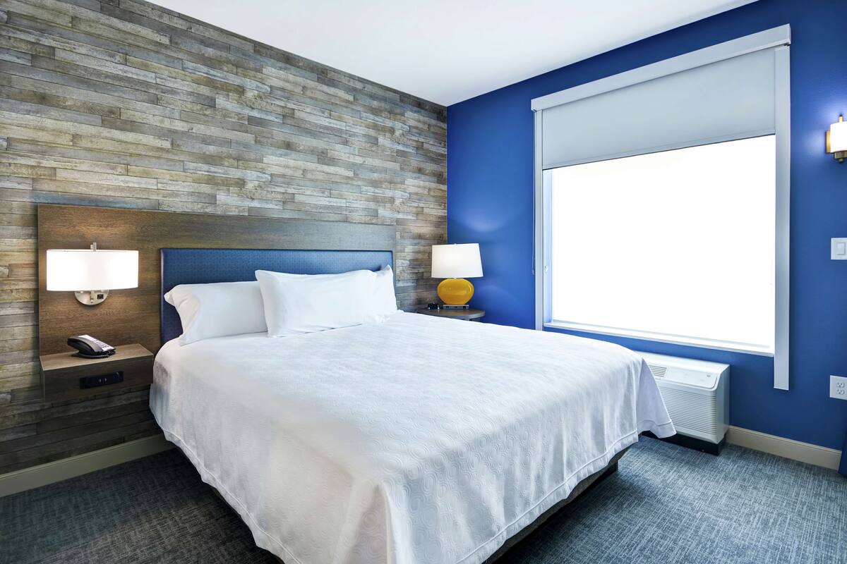 A hotel room with a large bed, wooden wall paneling, a window with blinds, and two bedside lamps.