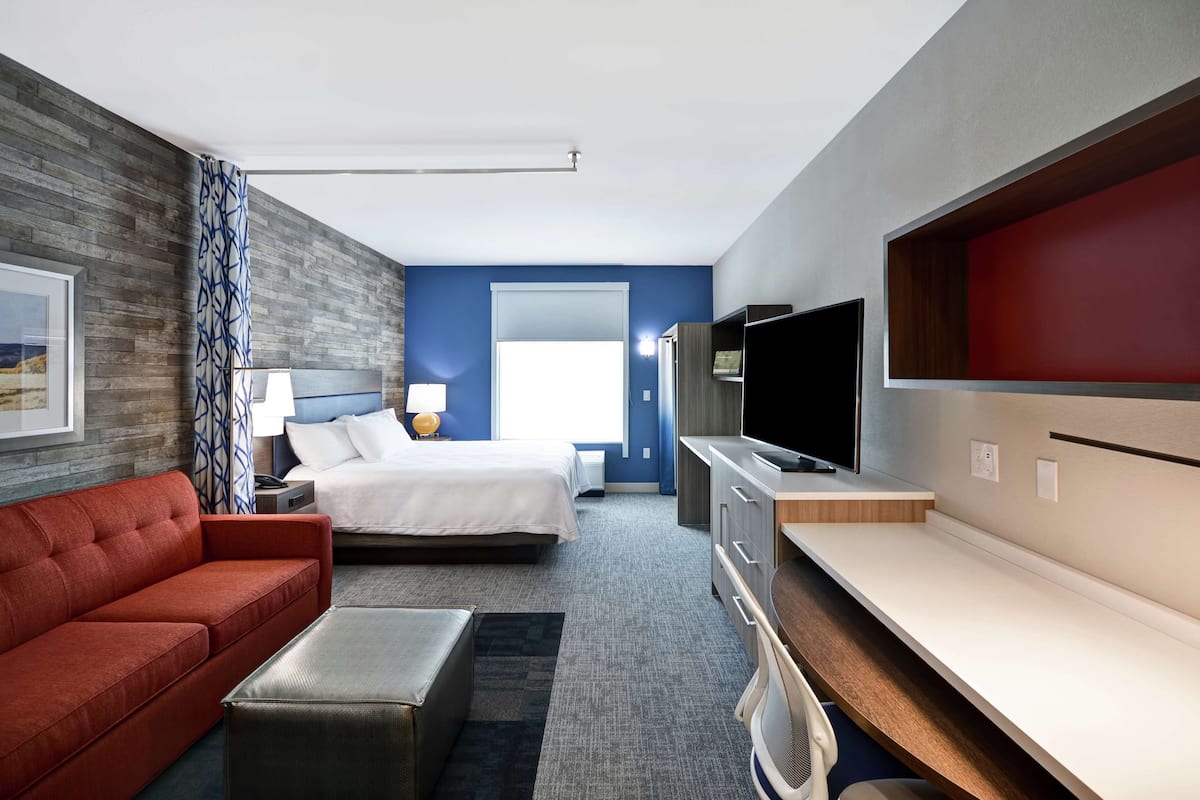 A hotel room with a bed, a red sofa, a TV, a desk, and a large window.