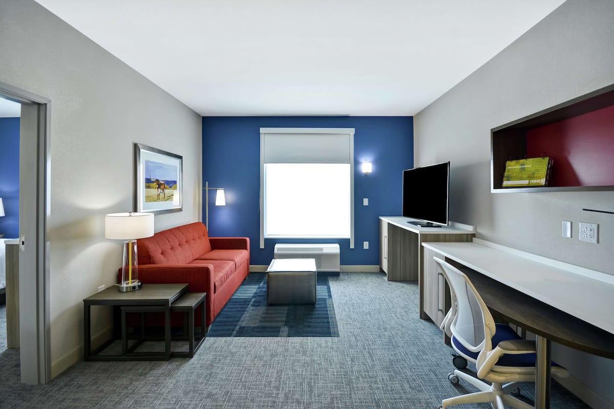 A hotel room with a red sofa, a small table with a lamp, a TV on a stand, a desk with a chair, and a large window.