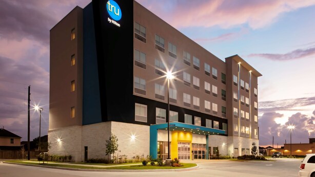 Un edificio moderno de hotel con un llamativo letrero 'tru by Hilton', varios pisos y una entrada bien iluminada.