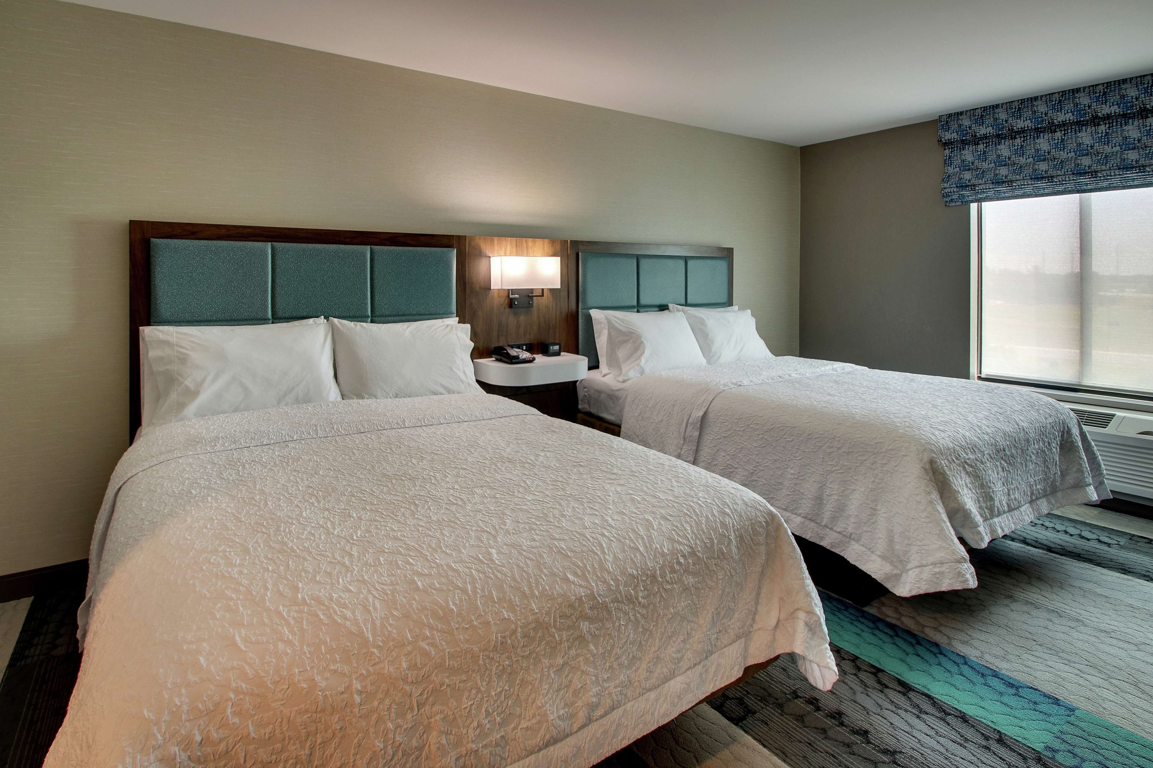 A hotel room with two beds, a nightstand, a lamp, and a large window.