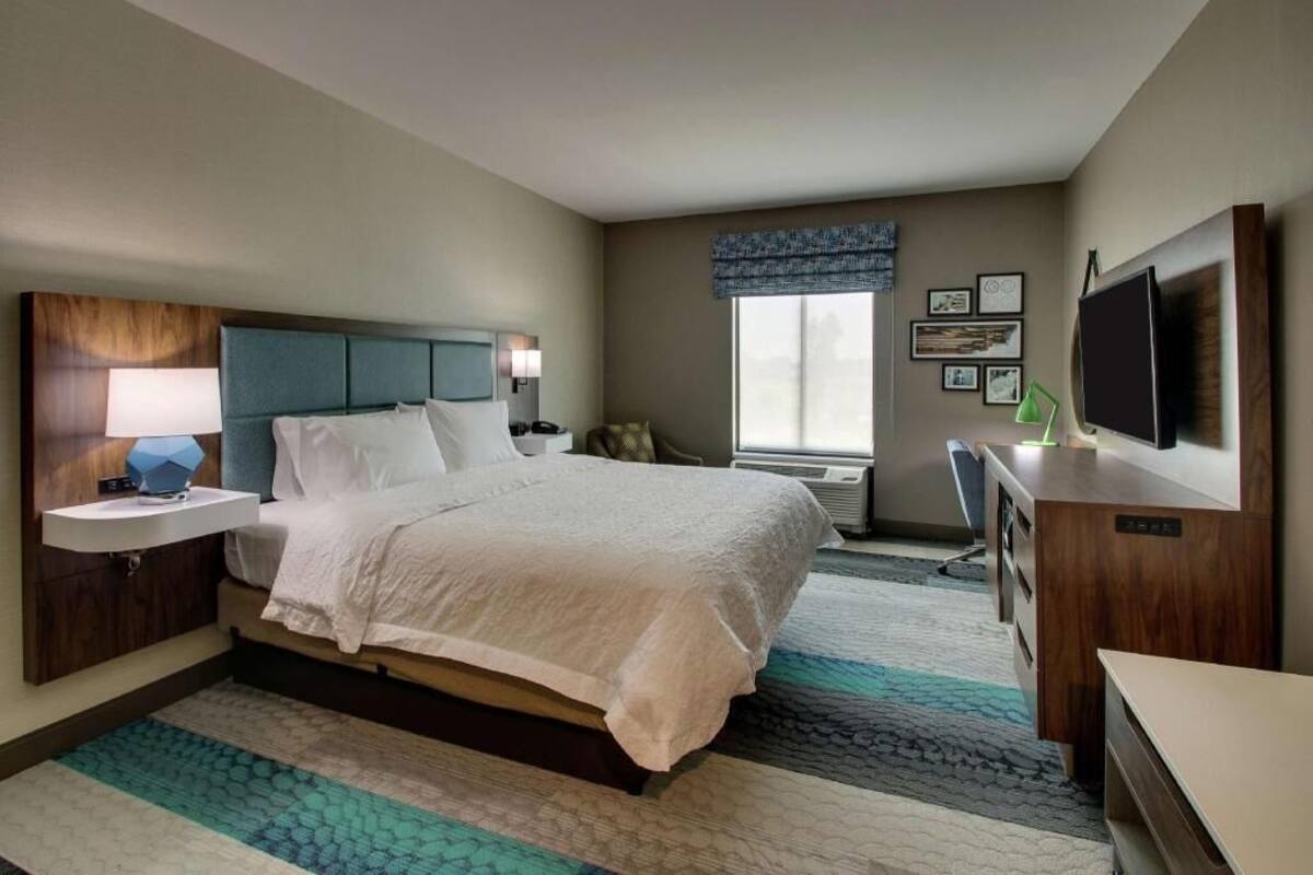 A hotel room with a bed, bedside table, lamp, TV, and a window with blinds.