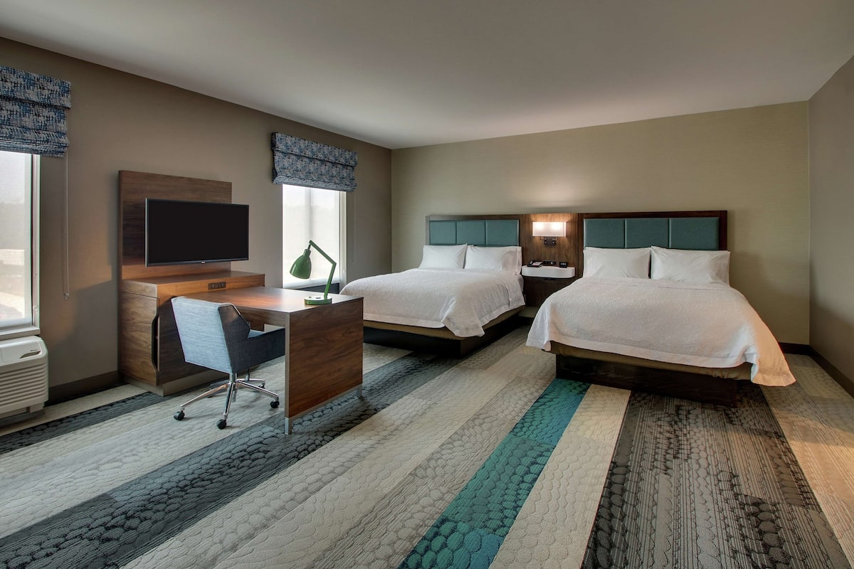 A hotel room with two beds, a desk, a TV, and a window with blinds.