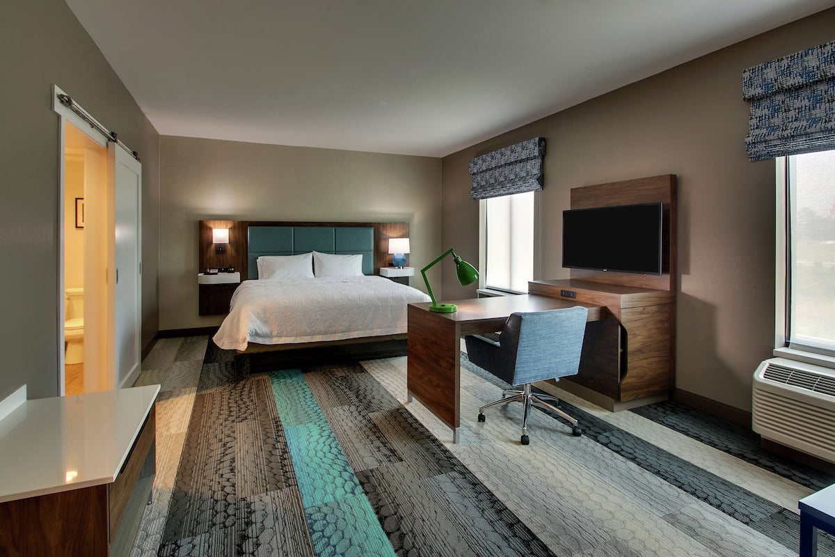 A hotel room with a bed, desk, chair, TV, and air conditioning unit.