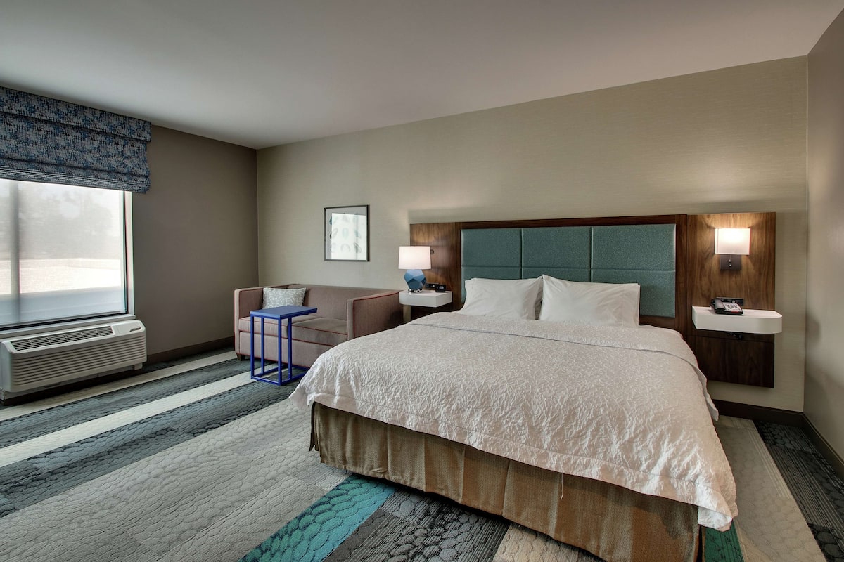 A hotel room with a large bed, a desk, a chair, a window with blinds, a wall-mounted lamp, and a bedside table with a phone.