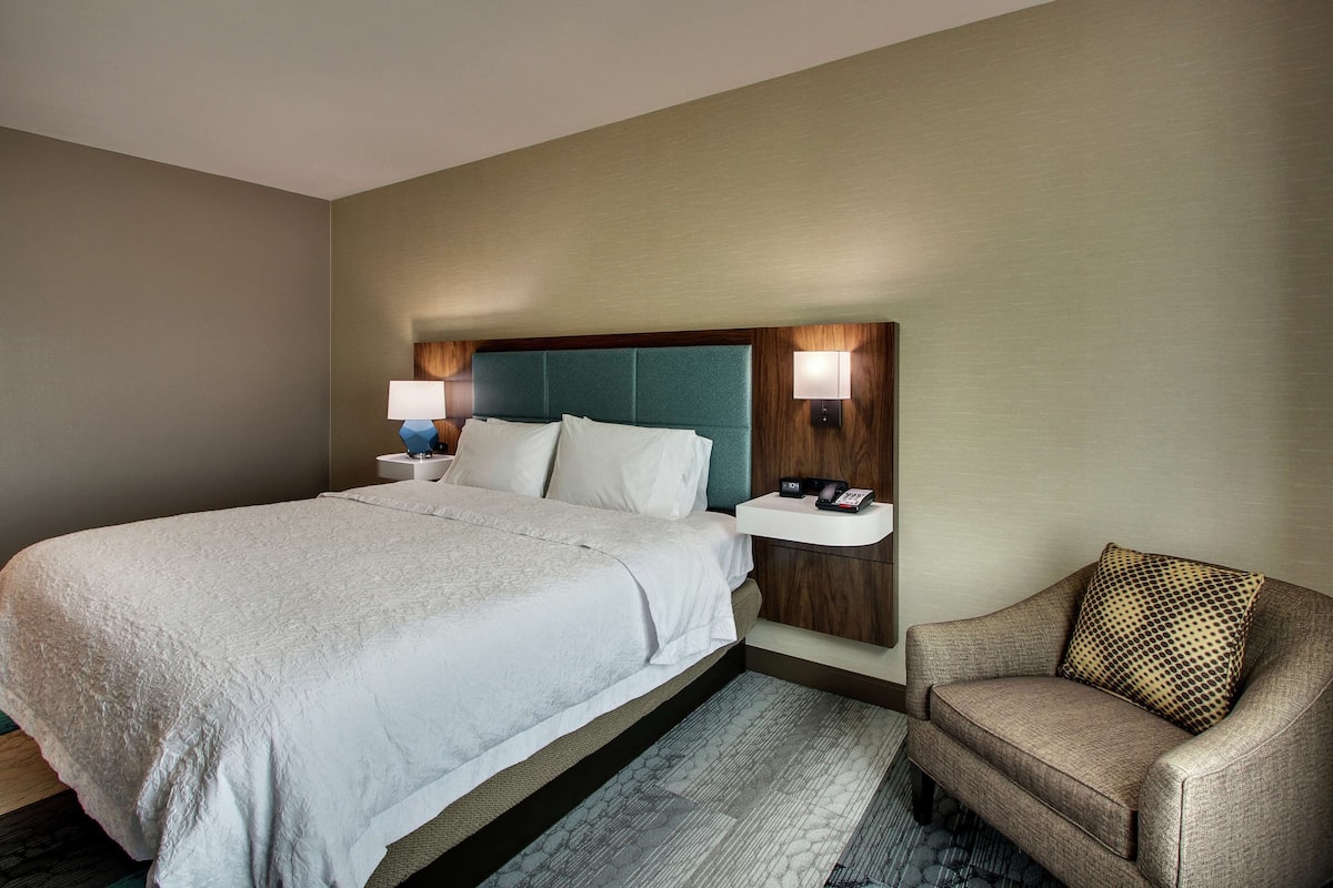 A hotel room with a large bed, a nightstand with a lamp, a chair, and a wall-mounted light.