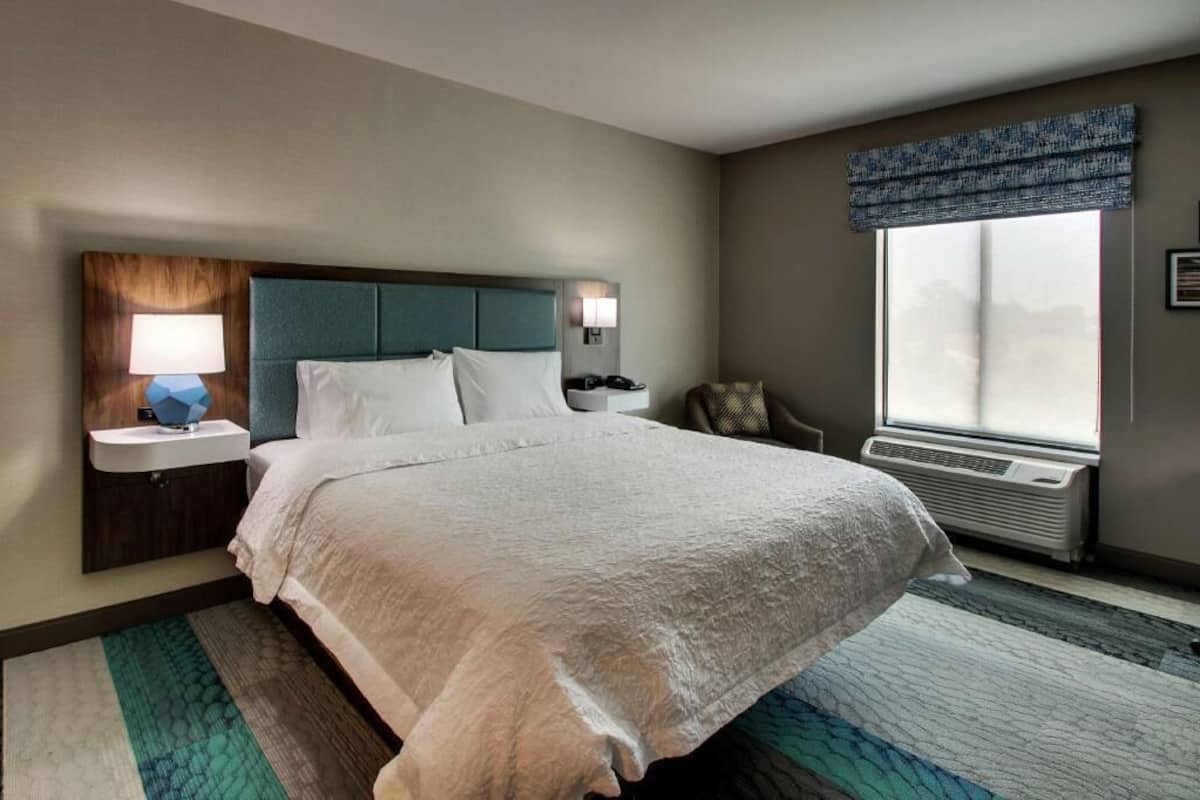 A hotel room with a large bed, bedside tables with lamps, a window with a view, and an air conditioning unit.