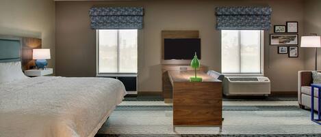 A hotel room with a large bed, a desk, a TV, and two windows with blue valances.