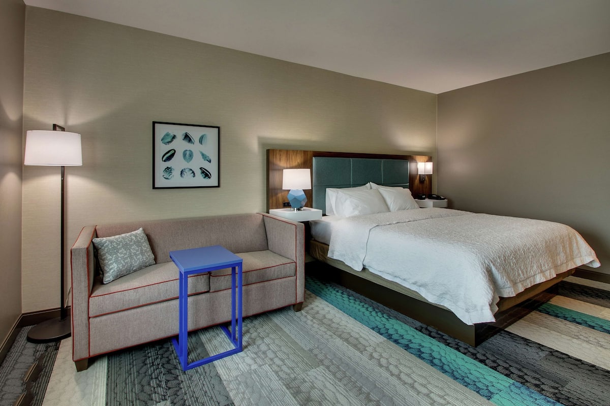 A hotel room with a bed, a sofa, a small table, a nightstand, a lamp, and a wall art piece.