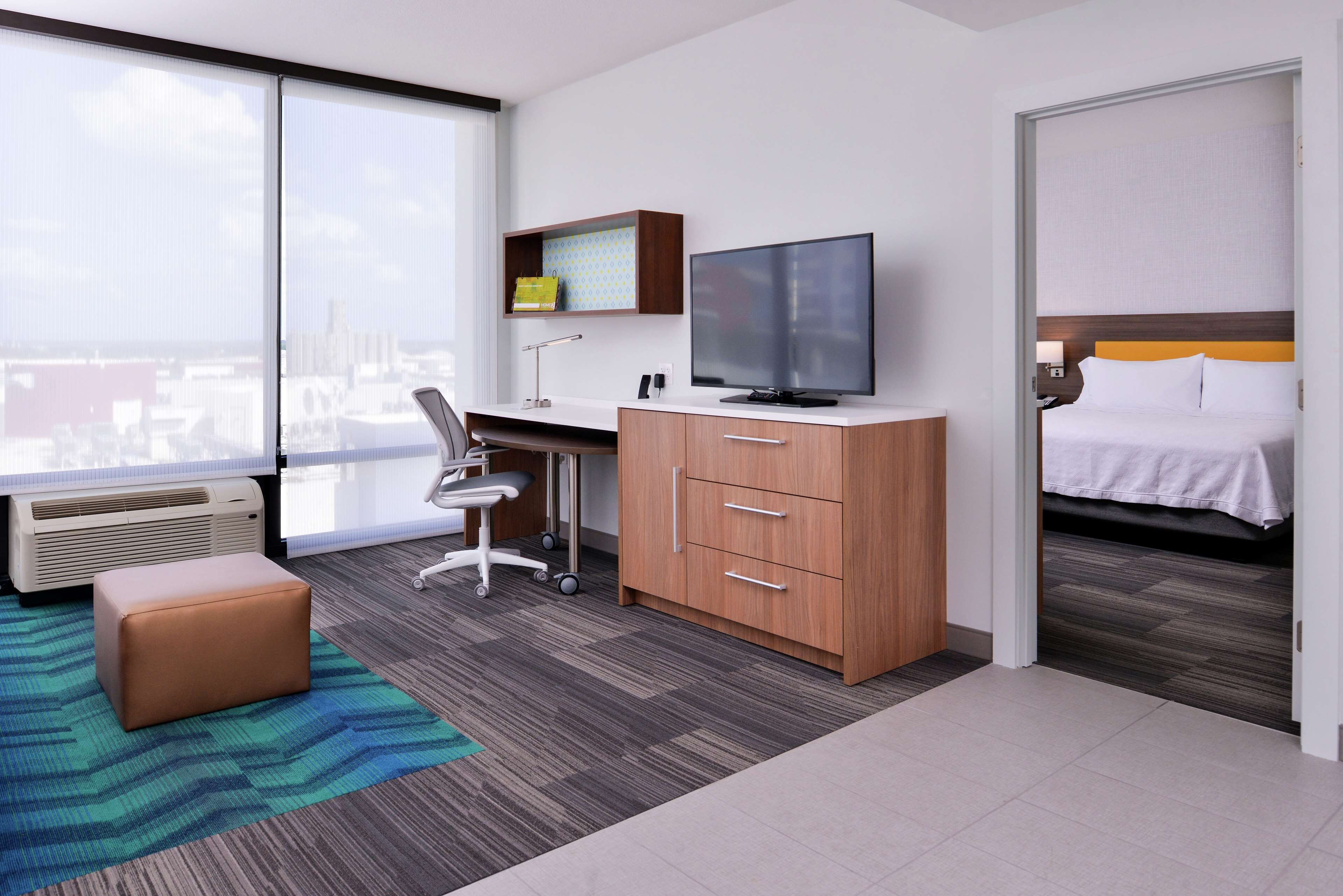 A hotel room with a large window, a desk with a computer, a television, a bed, and a small seating area.