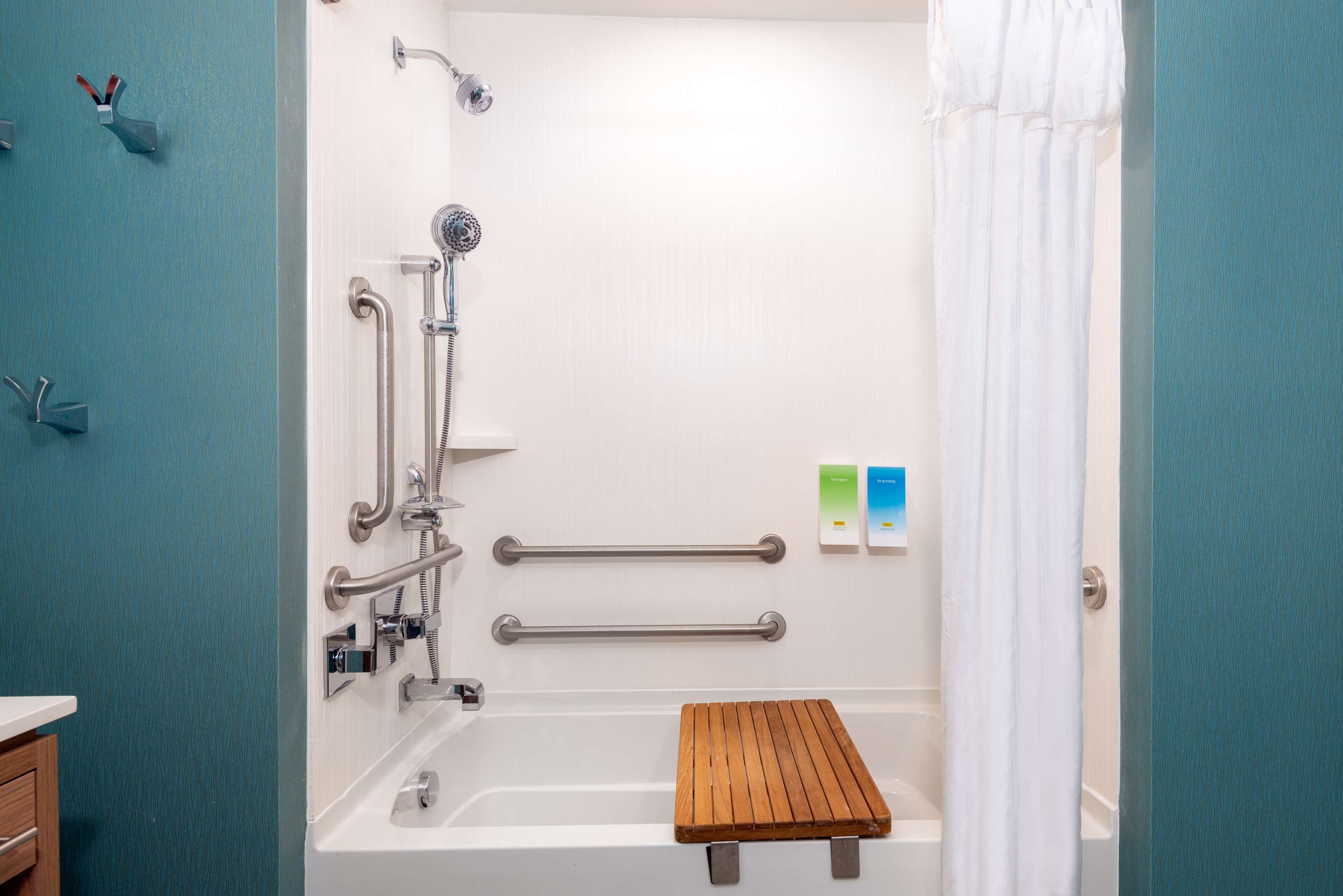 Une salle de bain avec une baignoire blanche, des barres d’appui, une pomme de douche et une étagère en bois.