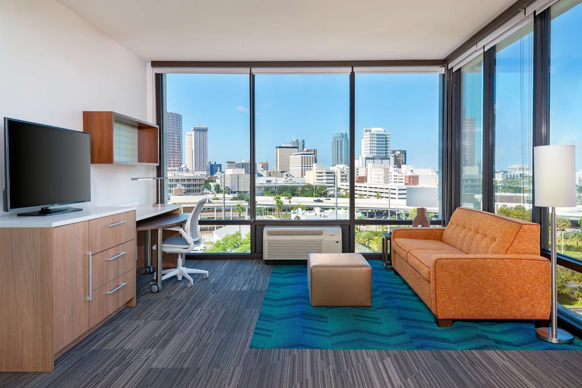 A modern hotel room with a large window, a flat-screen TV, a desk, a sofa, and a city view.