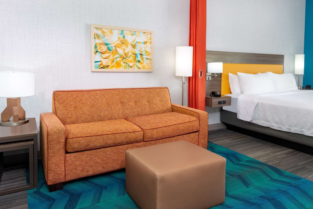 A hotel room with a sofa, a small ottoman, a bed with white linens, a nightstand with a lamp, a framed abstract painting, and a red curtain.