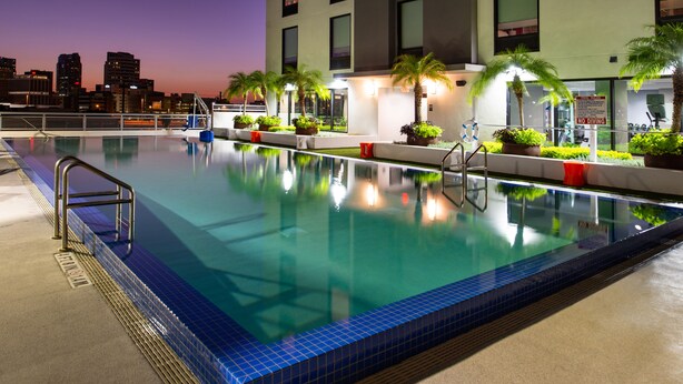 A swimming pool with a city skyline in the background, surrounded by palm trees and a building with a 'No Diving' sign.